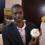 Daniel Kombat, CEO da Danikom Ghana Limited, produtora de manteiga de karité natural e sabonete preto africano, posa para fotos com produtos de manteiga de karité em um estande de exposição durante a conferência Shea 2026 em Accra, Gana, em 28 de abril de 2026. (Foto por Seth/Xinhua)